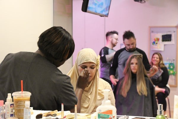 A teen has her makeup done. In the background another teen has her hair done.