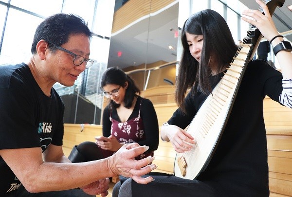 Three people seated in an atrium. One young woman is on her phone, while another woman and a man play a string instrument.