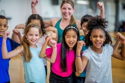 A group of elementary school students are posing with their teacher during gym class. They are all wearing athletic clothing, and flexing for the camera.