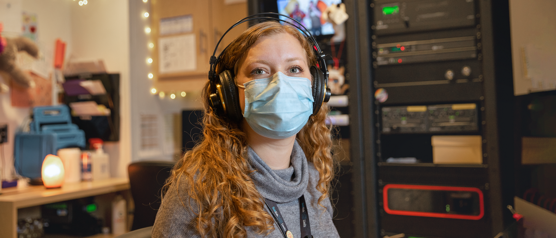 Staff member wearing headphones and a surgical mask sits at a desk with technical equipment in the background.