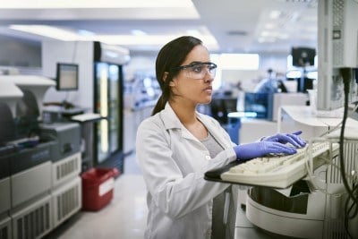 Shot of a woman in a lab coat and goggles using a computer while working in a laboratory