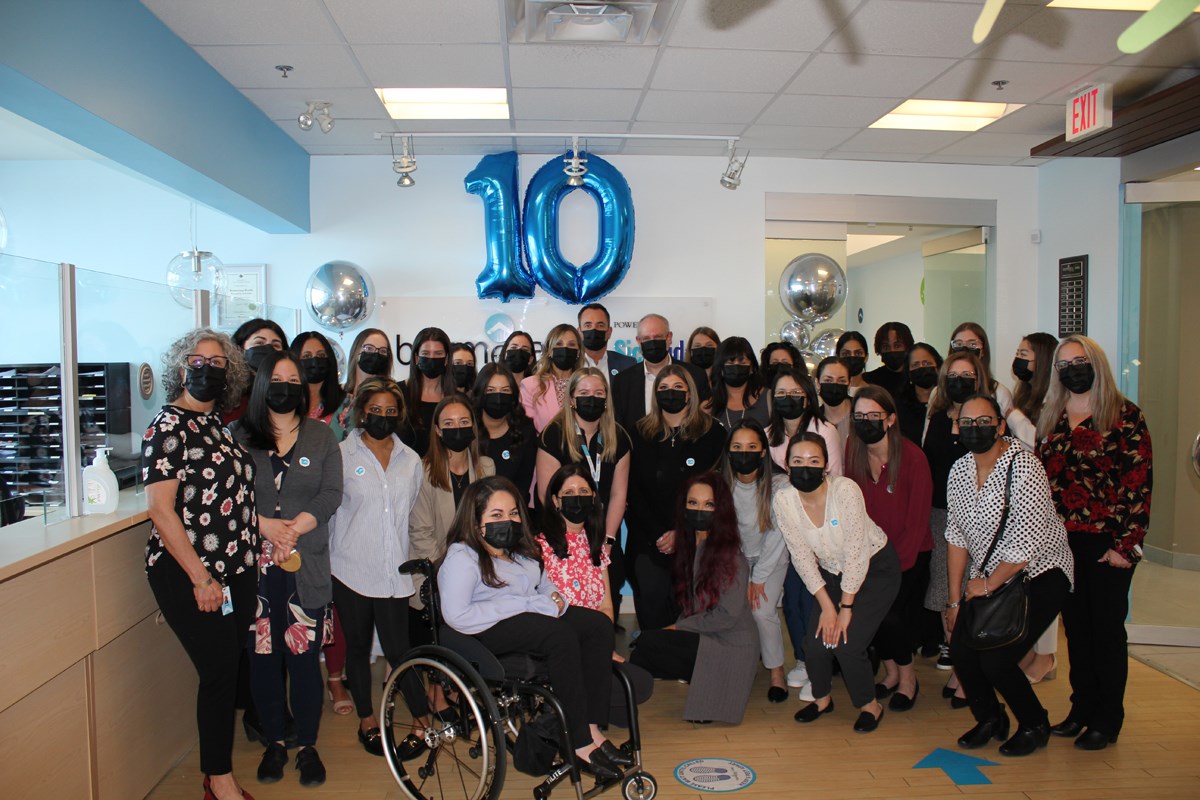 A large group of people standing together for a photo in masks. Behind them, there is a set of blue and silver balloons, with the blue balloons in the shape of the number 10.