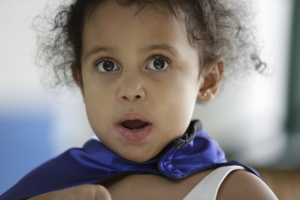 A young girl smiling timidly and wearing a super hero cape