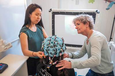 Drs. Margot Taylor and Julie Sato place an OPM powered helmet on a person's head.