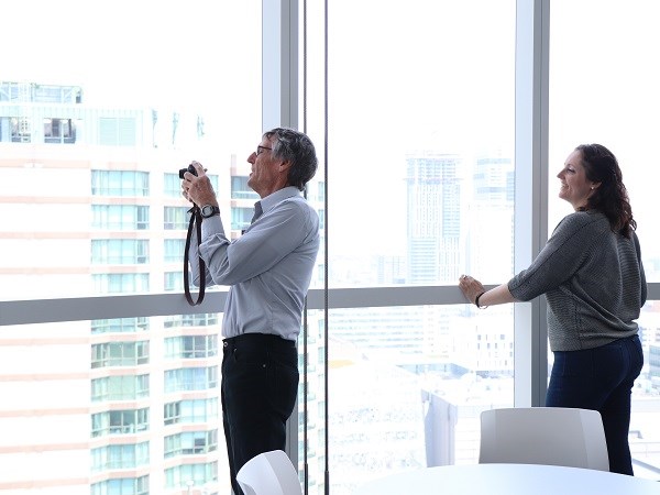 Two people look out the window of a very tall building, one holds a camera the other leans on a railing.