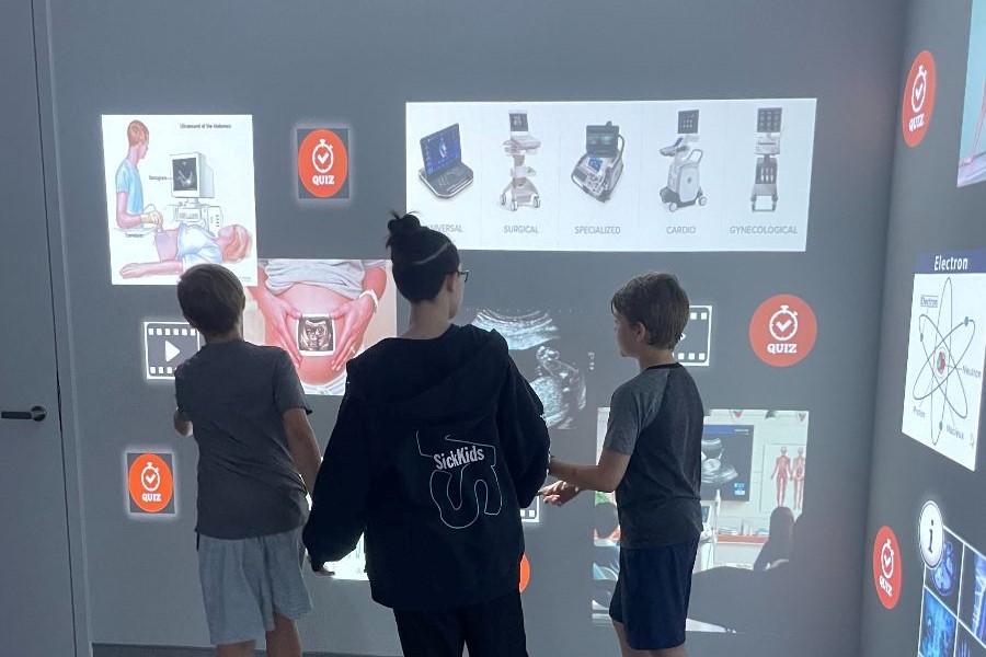 Three children engage with an interactive wall projection featuring medical images and quizzes in an educational setting.