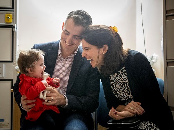 Baby sits on her father's lap. Her mother leans towards her. They are all smiling.