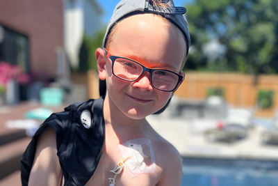 Owen Rose stands smiling outside a pool in his backyard, with medical tubes attached to his chest. 