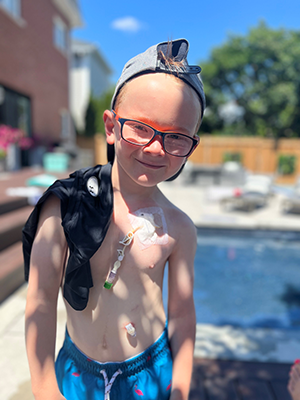Owen Rose stands smiling outside a pool in his backyard, with medical tubes attached to his chest.