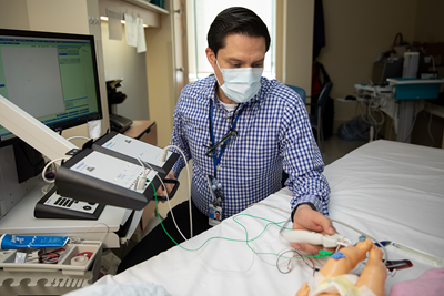 A man in a mask seated by a hospital bed and a computer monitor. He has a device in his hand that is hooked up to the computer and electrodes placed on a plastic infant model.