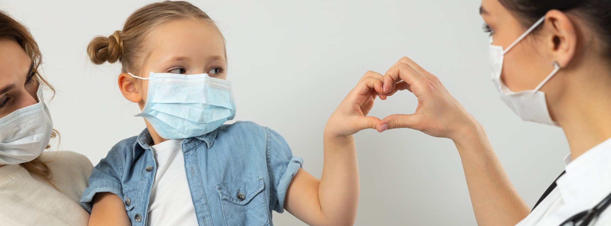 mother holding young girl who is making a hand-heart with her doctor