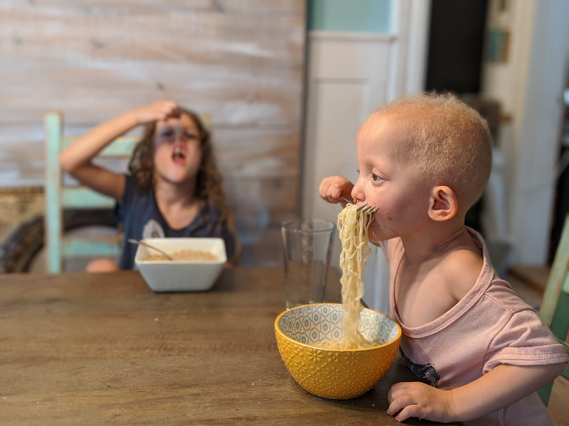 Two children sit at a table eating noodles.