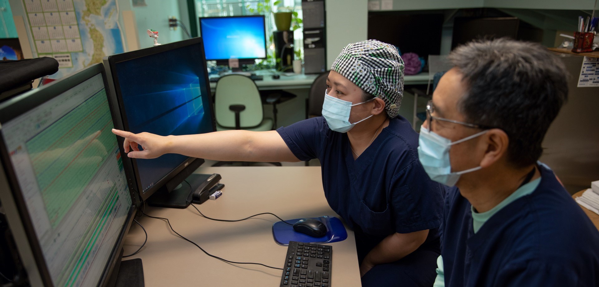 Paula Melendres pointing at a computer monitor displaying EEG signals. Hiroshi Otsubo is seated beside Paula and is observing the screen.