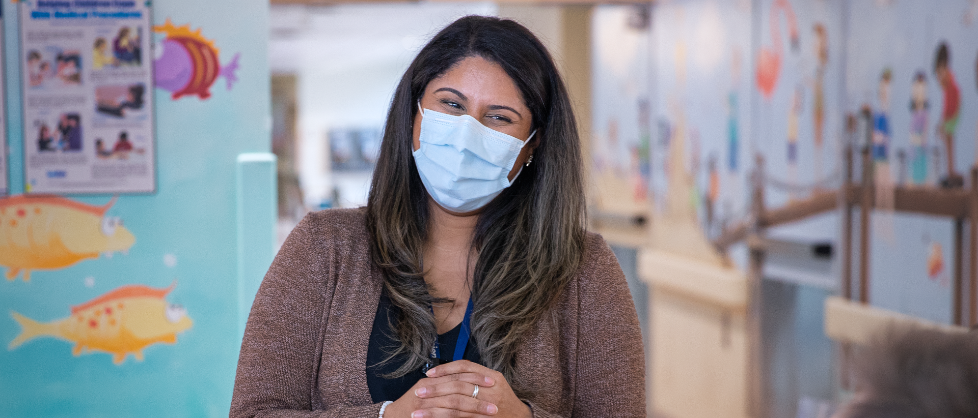 Person wearing a mask and smiling. They are standing in a clinical area with fish painted on the walls.