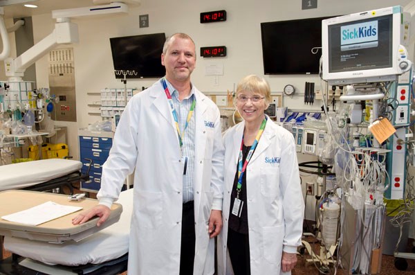 Two doctors stand together in an emergency department room filled with equipment. They wear lab coats.