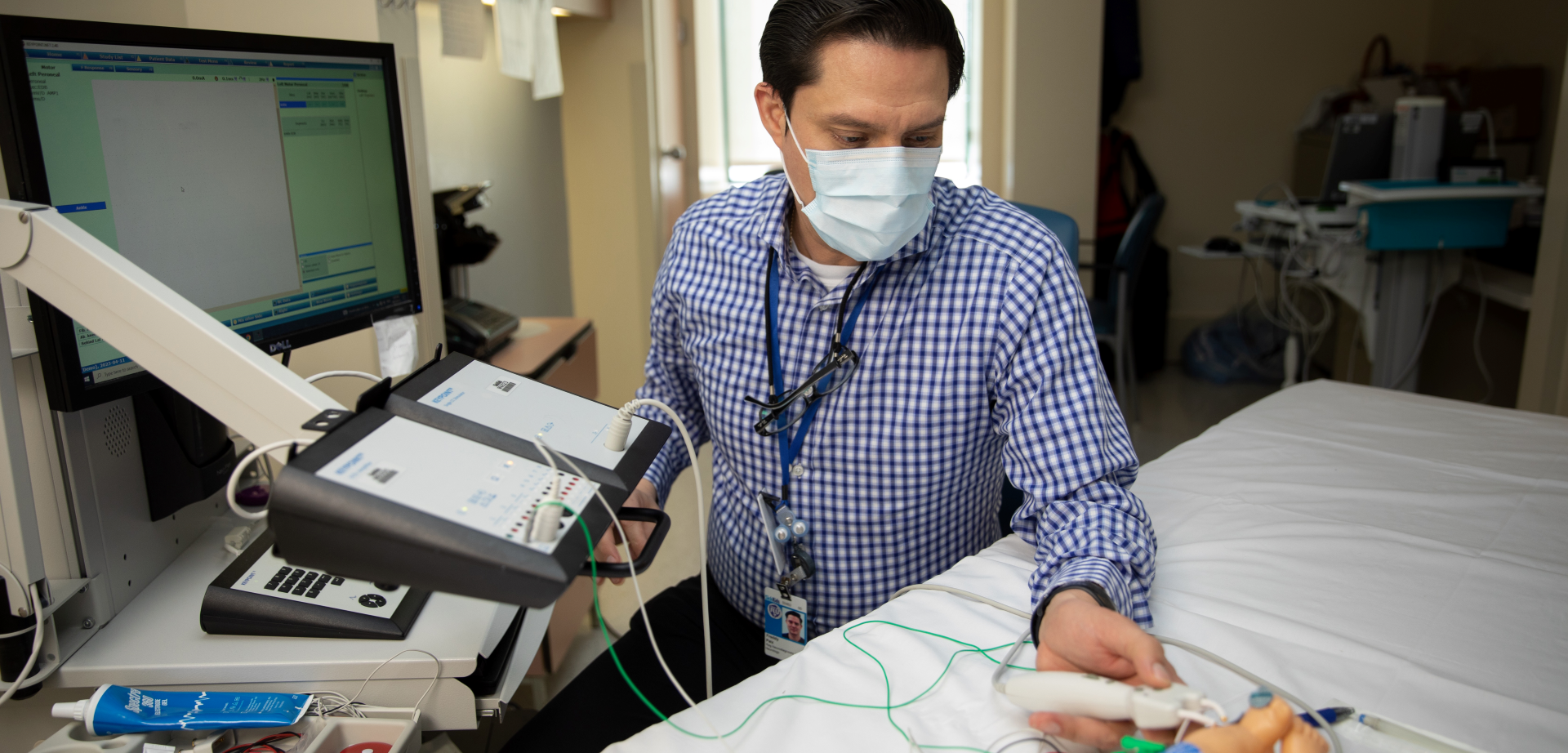 Freddy Paiz in a mask, seated by a hospital bed and a computer monitor. He has a device in his hand that is hooked up to the computer and electrodes placed on a plastic infant model.