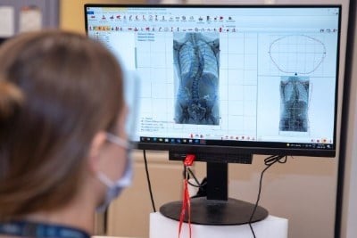 A person wearing a clear face shield and a surgical mask looks at a computer screen displaying black-and-white images of an X-ray of a person’s back. 