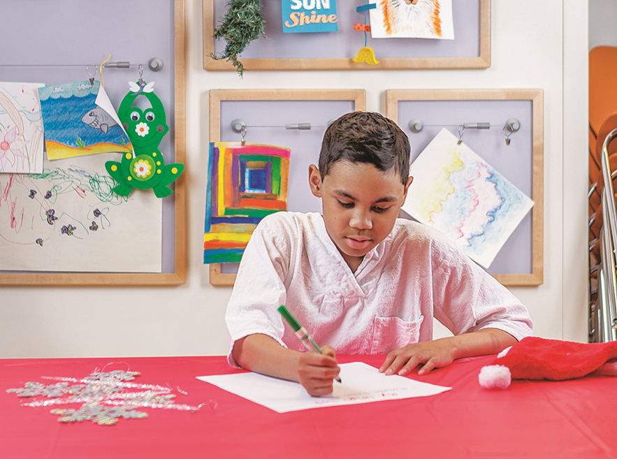 A child sits at a table drawing on white paper with a green marker, surrounded by craft items and colorful artwork on the wall.