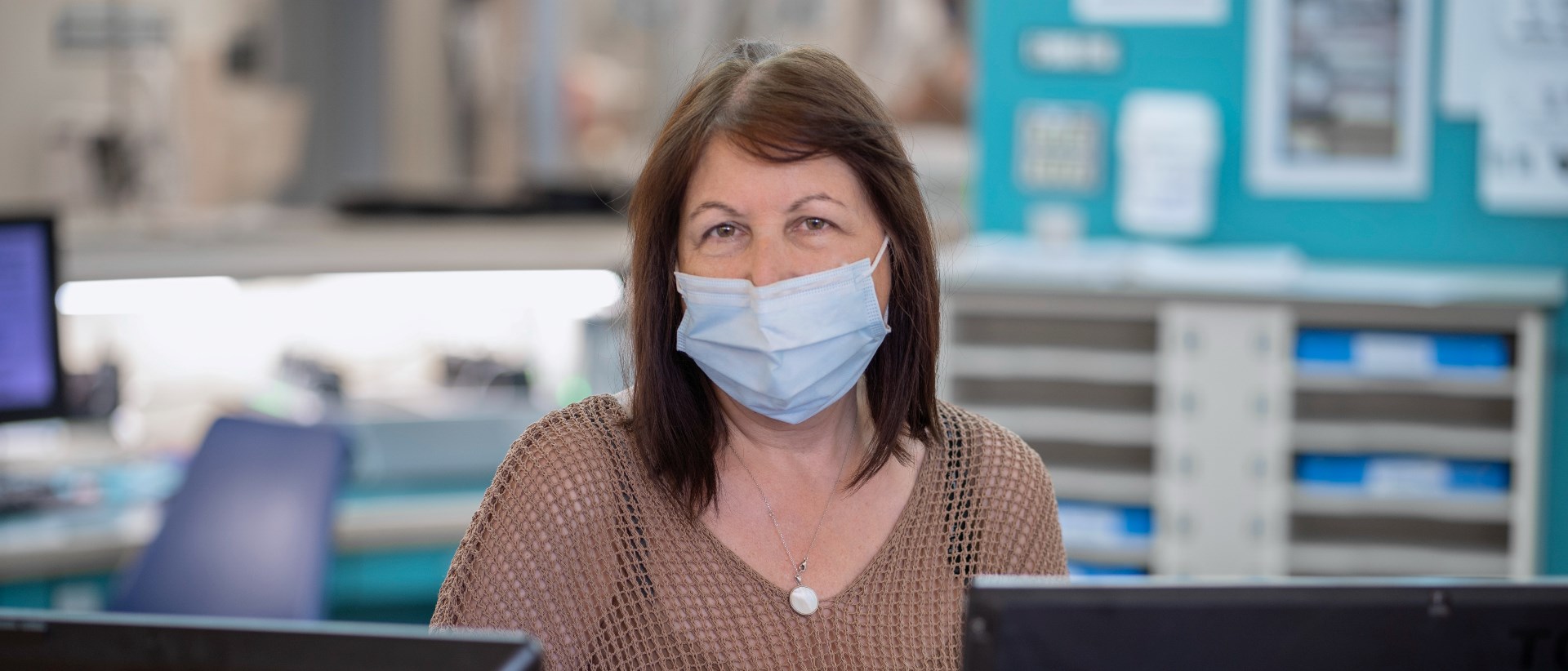 Rose Ancona in a mask sitting at a desk with two computer monitors.