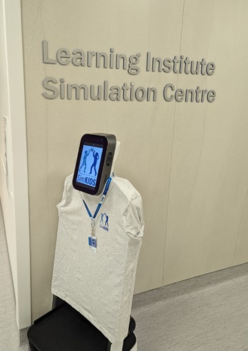 SimKIDS robot dressed in a branded t-shirt and lanyard in front of a wall that says "Learning Institute Simulation Centre"