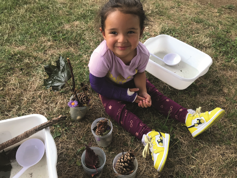 A girl seated on the grass with a collection of pinecones, leaves and sticks.