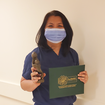 A woman wearing a mask and scrubs holds a small black statue and a green folder that says Daisy Award