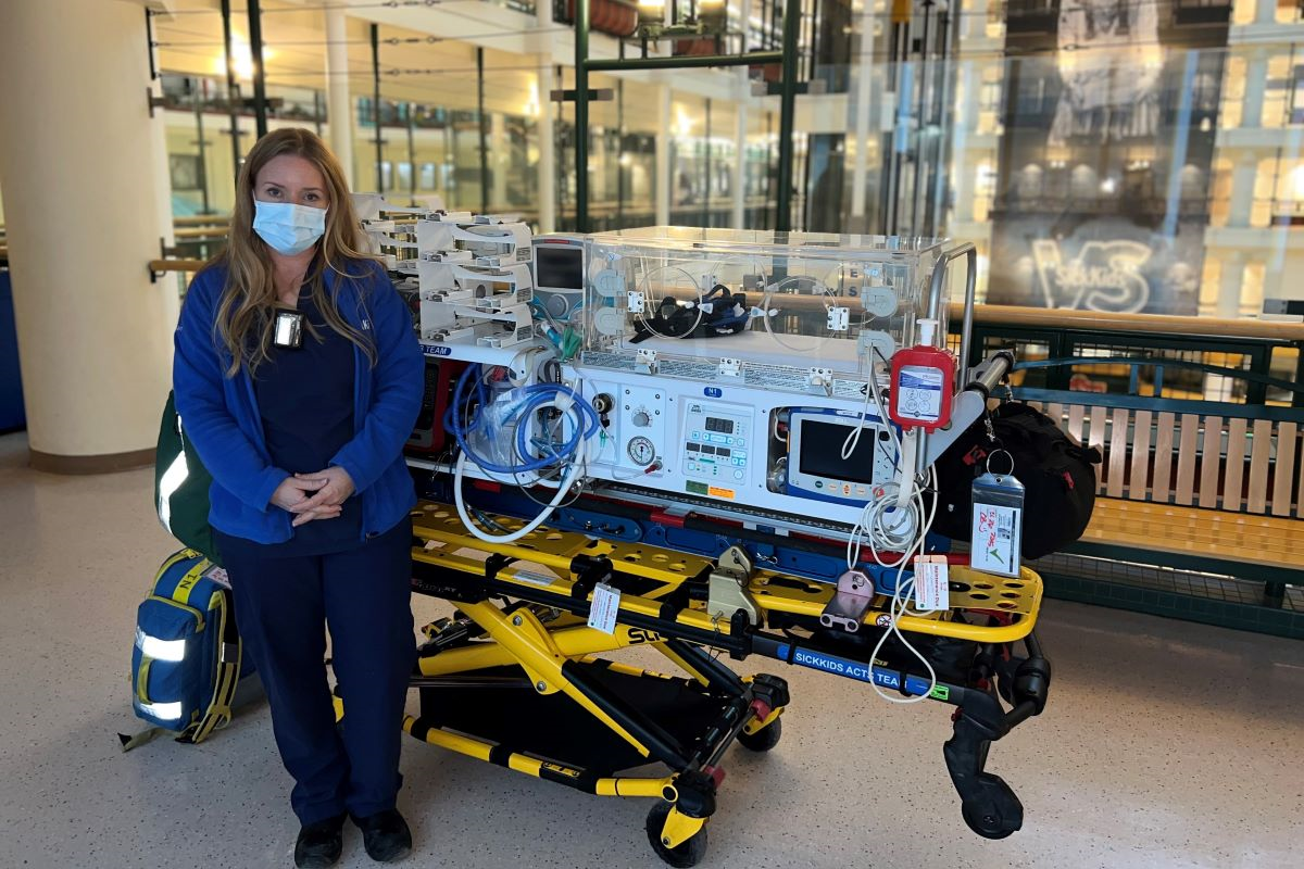 ACTS Registered Nurse Clinician Debbie McFarlane stands next to a mobile NICU incubator wearing a mask.