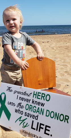 A toddler stands beside a wooden chair on the beach. A sign leans against the chair, reading "The greatest hero I never knew was the organ donor who saved my life."