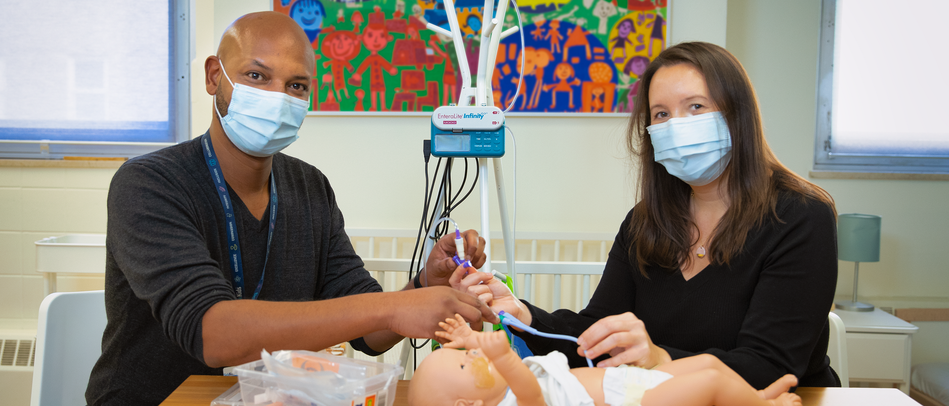 Two people seated at a table, demonstrating how to insert a nasogastric tube into a simulation doll.