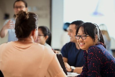 A woman in glasses smiling and chatting with another woman sitting beside her, with other people smiling and talking in the background.