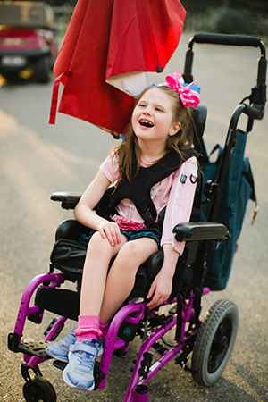 Young girl with Rett syndrome smiling in a wheelchair.
