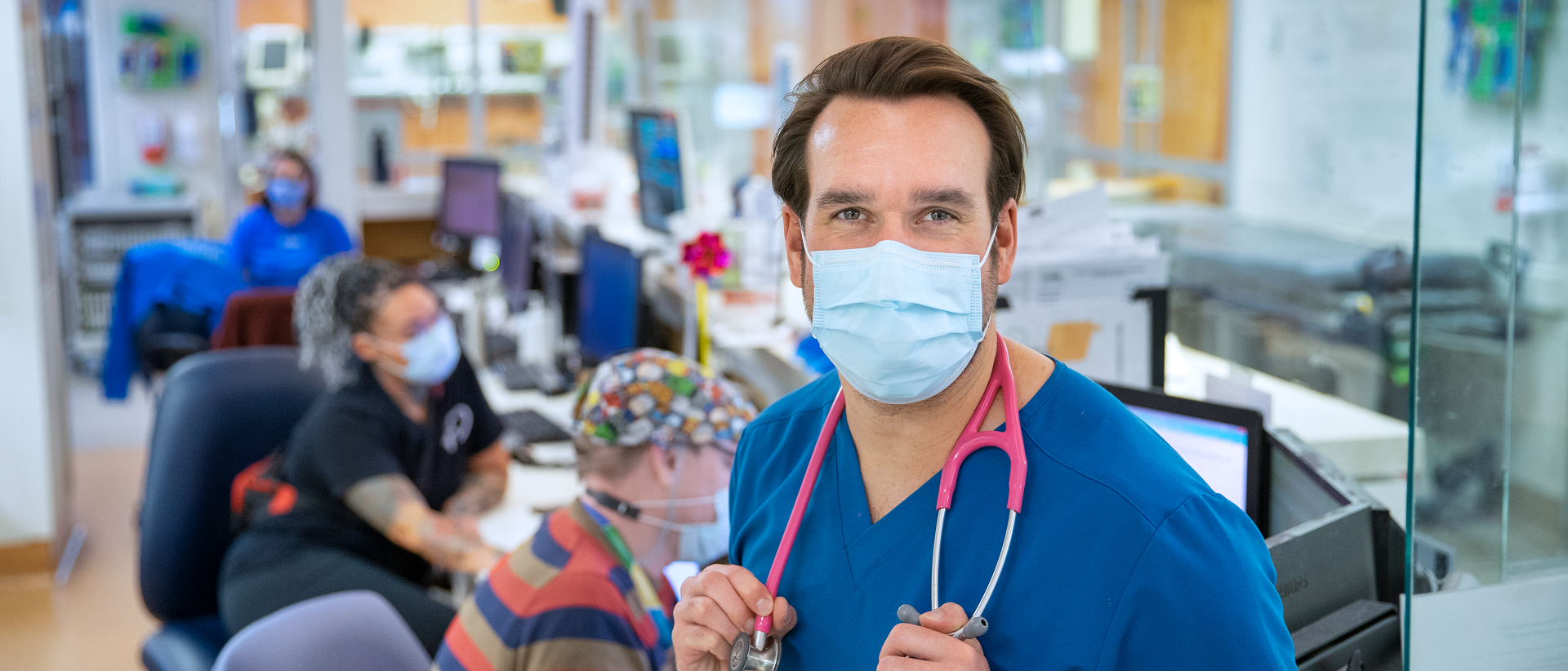 Man wearing scrubs, a surgical mask and a stethoscope around his neck stands in a clinical area.
