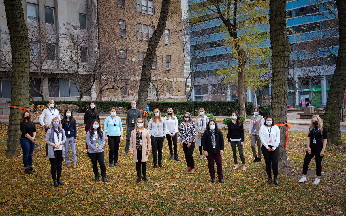 People stand on a lawn with trees and a building in the background. They are all wearing masks and distanced from each other.