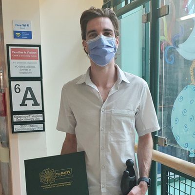 A man wearing a mask holds a small black statue and a folder that reads Daisy Award