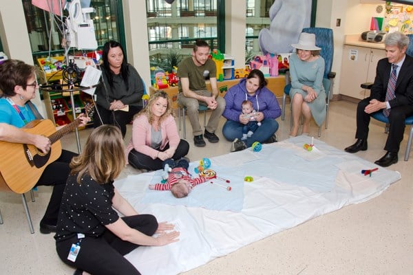 Group of adults and babies seated on the floor and low chairs. One person holds a guitar.