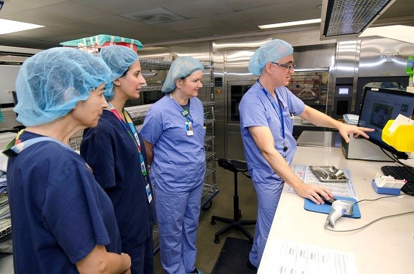 One staff member points at a computer and operates the mouse as others look on from behind him. They are all wearing scrubs and hairnets.