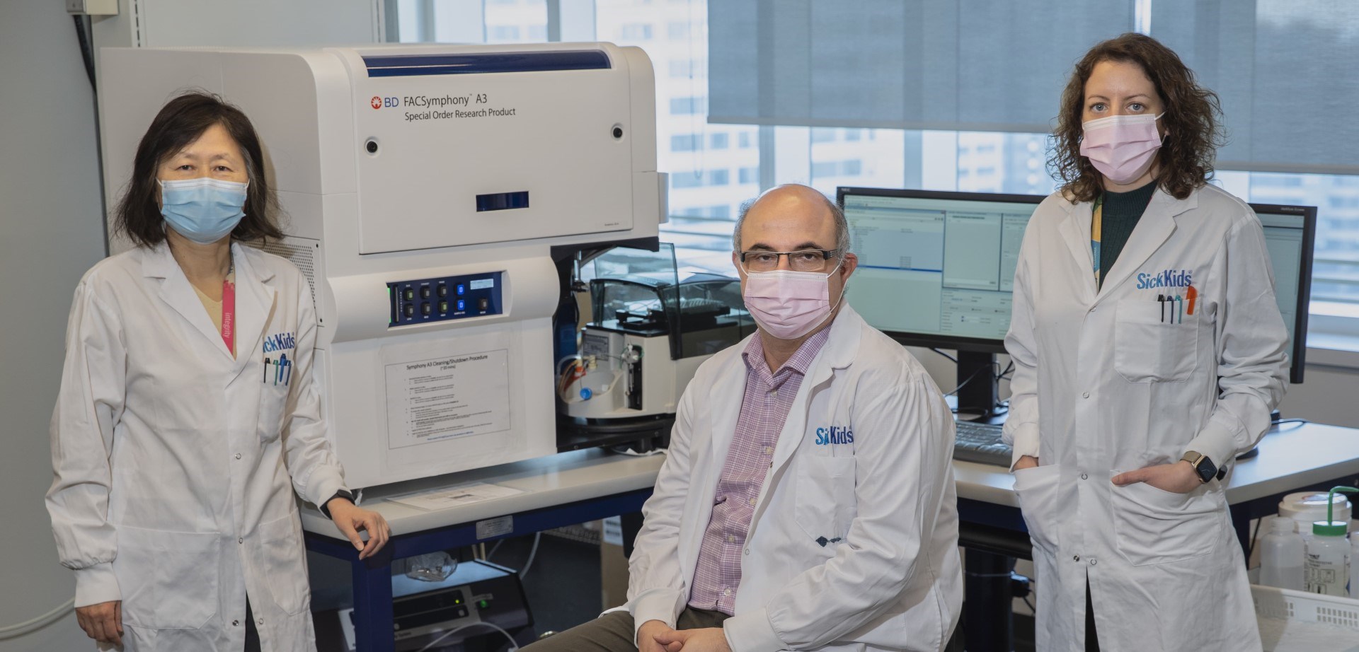 (left to right) Sheyun Zhao, John Moniakis, and Emily Reddy in masks and lab coats. There is a computer and lab equipment behind them.