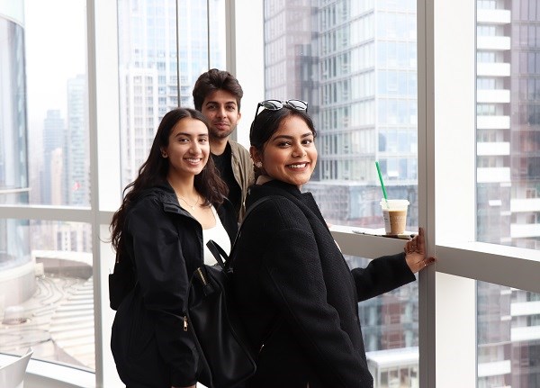 Three people smile at the camera with a window and skyscrapers seen behind them.