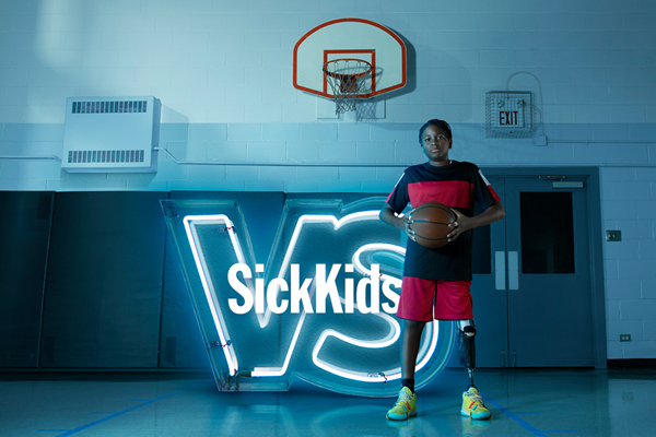 A boy with a prosthetic leg holding a basketball in his hands. He is standing in front of a neon sign of the SickKids VS campaign logo under a basketball net in a school gym.