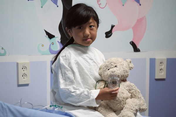 Child wearing hospital gown holds stuffed teddy bear.