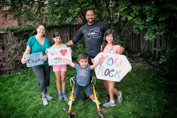 A woman, man and three children stand in a backyard while holding up handmade signs that say "SickKids Thanks", "We love SickKids", and "You rock". The man is wearing a SickKids VS t-shirt. A boy with an assistive walking device stands in the centre with his arms raised in the air.