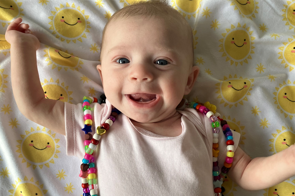 An infant lying on a sun-patterned blanket. The infant is wear bravery beads and smiling up at the camera.