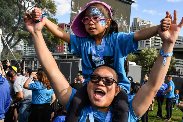 Child on an adult's shoulders. They are wearing matching shirts, holding hands and cheering.