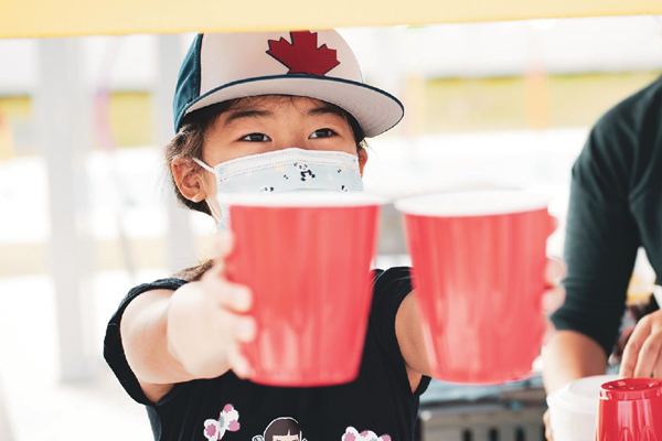 Youth wearing a baseball hat and face mask holds two cups in their outstretched arms.