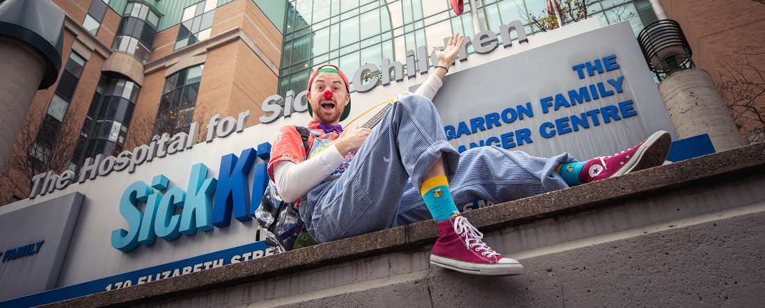 Clown sits outside the sign for The Hospital for Sick Children, smiling and waving.