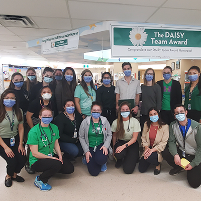 A group of 19 staff in masks huddle together on a hospital unit under a hanging banner that reads The Daisy team award