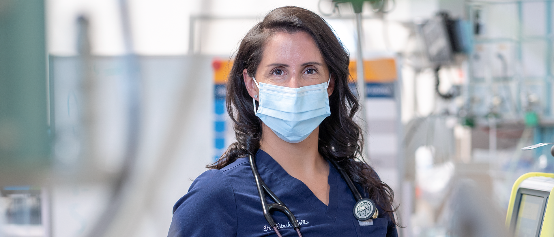 Dr. Natasha Collia in a mask with a stethoscope around her neck. There is a variety of hospital equipment out of focus in the background.