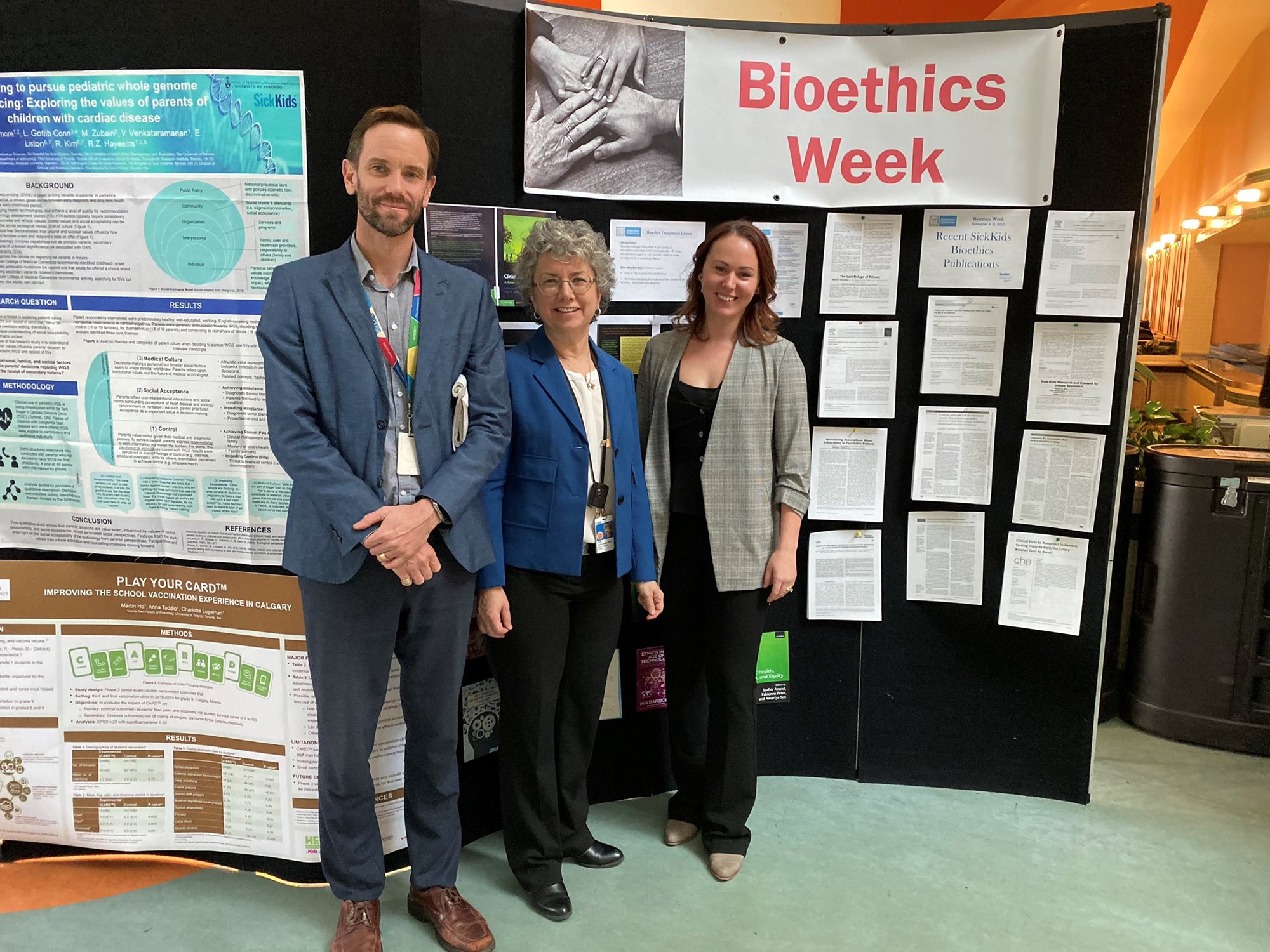 Three staff stand together in front of a poster display with a banner that reads Bioethics Week in the background.