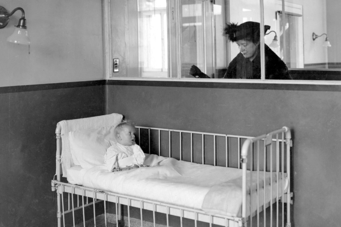 Black and white photo of infant in hospital crib with person watching through window glass.