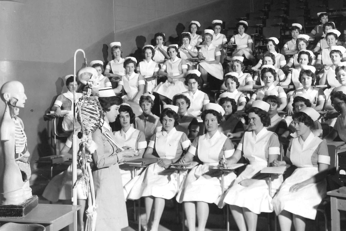 This black and white image shows nursing students in uniform with white caps in a classroom. An instructor stands near anatomical models (skeleton and torso) while teaching.